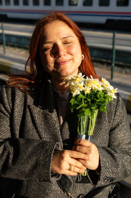 A woman with red hair holding a bouquet of fresh white and yellow daisies with green foliage, wrapped in clear cellophane and tied with a ribbon, standing outdoors near a railway track. The flowers ar