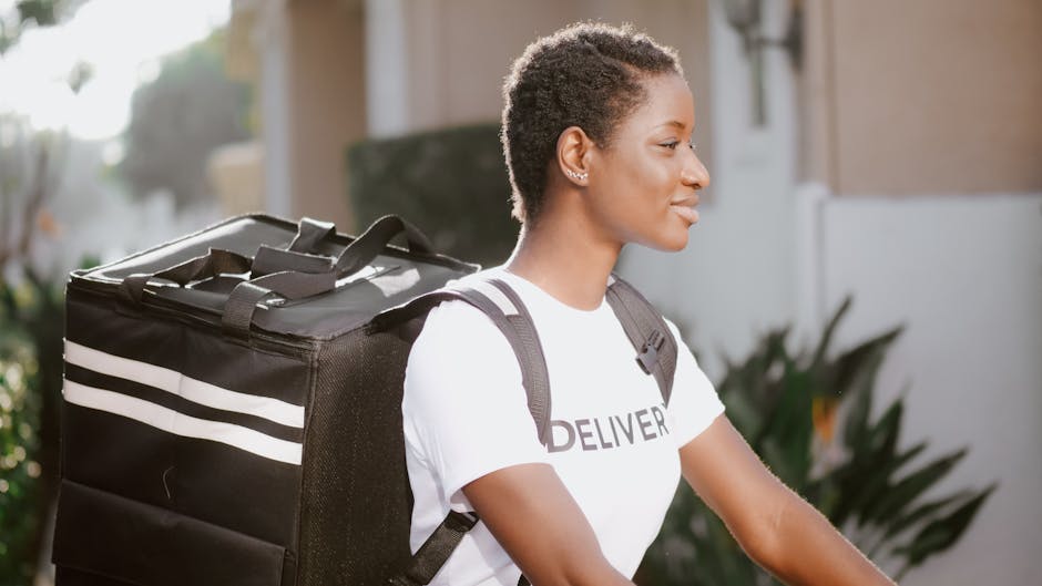 A smiling woman with short curly hair and medium-dark skin tone, dressed in a white T-shirt with 'DELIVERY' printed on it, carries a large black insulated delivery bag with white stripes over her shou