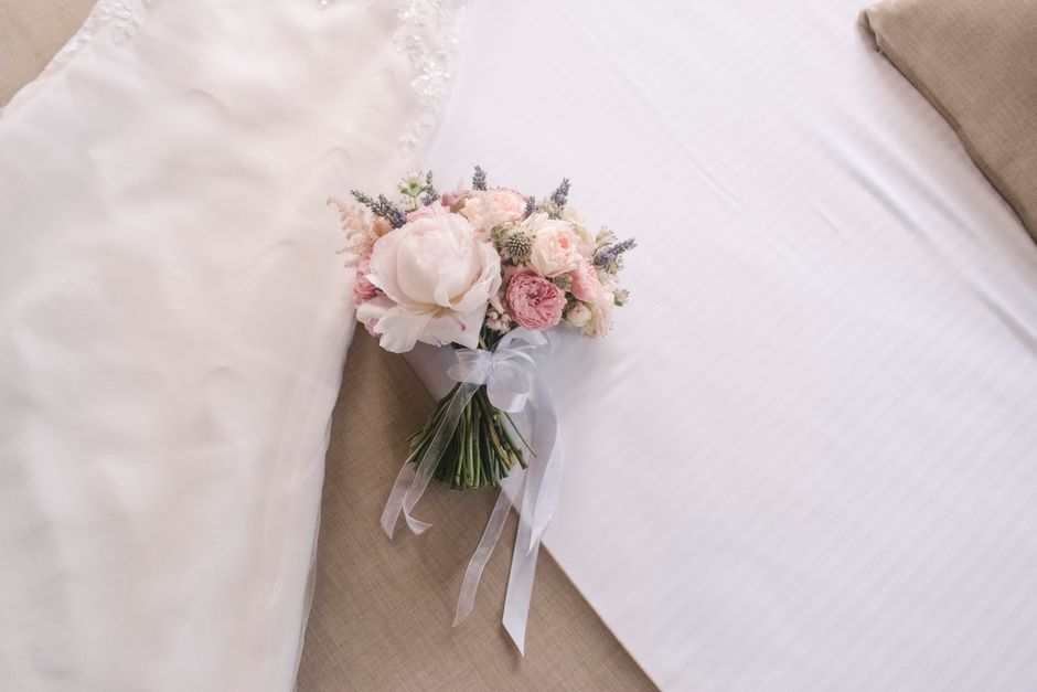 A bouquet of fresh flowers resting on a bed, featuring large white peonies, soft pink roses, and sprigs of lavender, arranged in a small white vase wrapped with a delicate white ribbon. The bouquet is