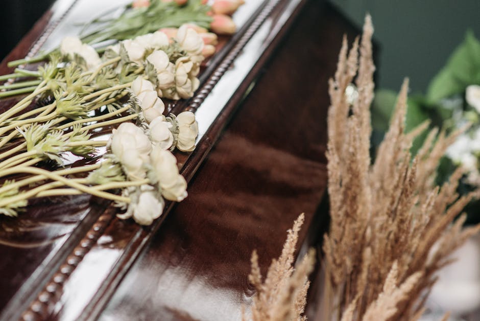 A close-up of a floral arrangement resting on a polished wooden surface, featuring delicate white roses with tightly closed petals and pale green stems, arranged in a loose, natural style. To the righ